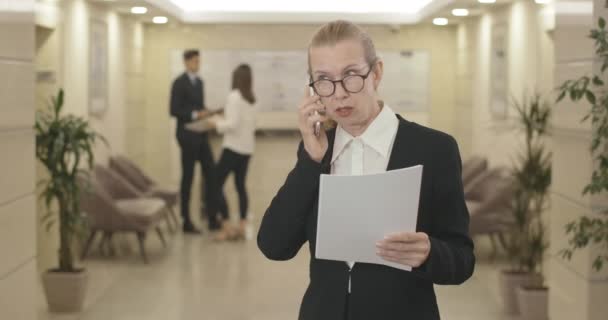 Впевнена бізнесменка середнього віку в окулярах обговорює проект по телефону, тримаючи в руках документ. Портрет елегантної кавказької жінки, що стоїть у діловому центрі і розмовляє. Фільм 4k ProRes HQ. — стокове відео