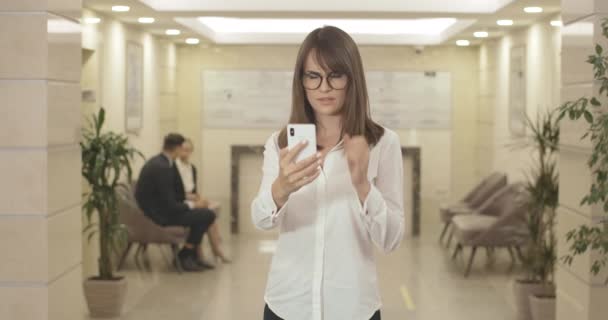 Plan médian d'une jeune femme d'affaires portant des lunettes ayant des problèmes de vision. Portrait d'une femme caucasienne stressée et confuse essayant de lire sur l'écran du smartphone dans le hall du centre d'affaires. Siège social Cinema 4k ProRes. — Video