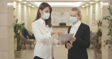 Maskeli genç ve orta yaşlı kadınlar ofiste projeyi tartışıyorlar. İş merkezinde Covid-19 salgınında çalışan ciddi iş adamlarının portresi. Coronavirus yaşam tarzı. Sinema 4k ProRes Merkezi.