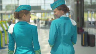 Havaalanındaki çıkışa doğru yürüyen ve konuşan iki pozitif hostesin arka görüntüsü. Kamera, eve dönen profesyonel uçuş görevlilerini izliyor. Kafkas kadınlarının havacılık sektöründe çalışırken orta pozu..