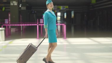 Pozitif gülümseyen hostes havaalanı terminalinde çantasıyla durup el sallıyor ve yürüyor. İnce, kendine güvenen beyaz hostes portresi. Geldikten sonra veya ayrılmadan önce..