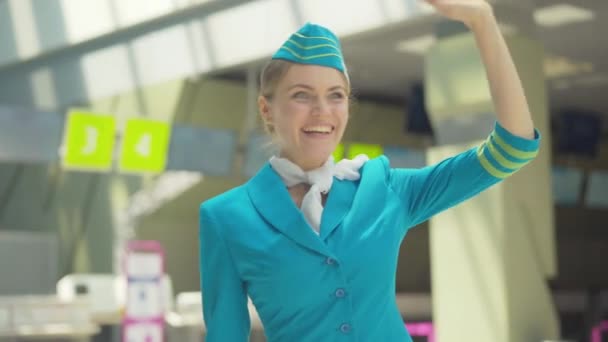 空港で手を振って魅力的な白人スチュワーデスの肖像画。到着後に誰かに笑顔の美しい若い女性。青い制服を着た自信のある女性客室乗務員出発を待っています. — ストック動画