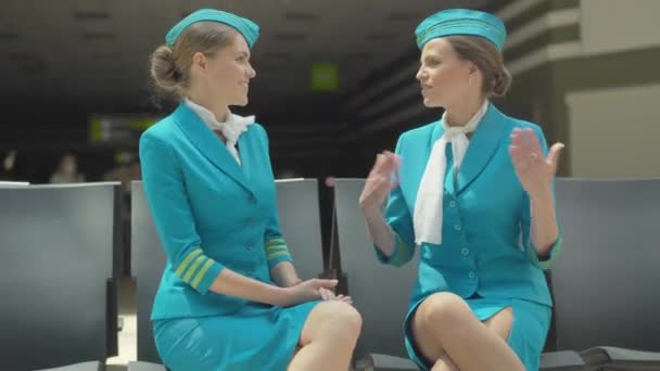 Portré két karcsú, gyönyörű kaukázusi nőről stewardess egyenruhában, akik a reptéren ülnek és beszélgetnek. Fiatal, magabiztos utaskísérők várják az indulást. Szakmai csevegés zárt térben. — Stock videók