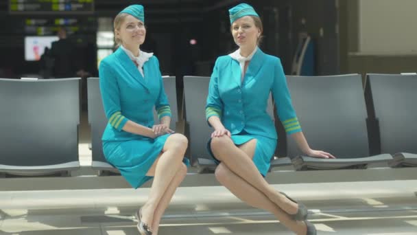 Amplia toma de dos bellas azafatas sentadas en la terminal del aeropuerto y hablando. Retrato de hermosas azafatas caucásicas confiadas esperando la salida. Tripulación profesional en uniforme azul. — Vídeo de stock