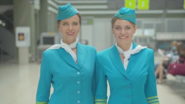 Mavi üniformalı ve kameraya gülümseyen iki beyaz hostesin orta pozu. Gitmeden önce havaalanında poz veren profesyonel uçuş görevlilerinin portresi.