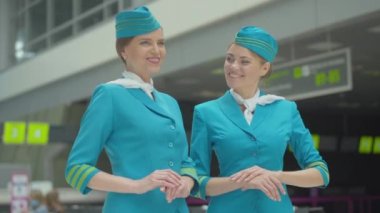 Kendine güvenen profesyonel hostesler sohbet ediyor, kameraya dönüyor ve gülümsüyor. Havaalanında poz veren güzel beyaz kadın portresi. Terminalde kalkış için bekleyen bayan uçuş görevlileri..