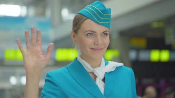 Közelkép egy mosolygós, gyönyörű, fehér nőről stewardess egyenruhában, aki félrenéz és integet. Pozitív magabiztos profi légikísérő áll a repülőtéri terminálon. Turisztikai koncepció. — Stock videók