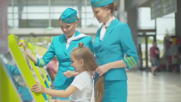 Positiva professionella flygvärdinnor hjälper söt liten flicka med terminal på flygplatsen. Sidovy av vackra vita barn att göra betalning stöds av leende flygvärdinnor. Medföljande, turism. — Stockvideo