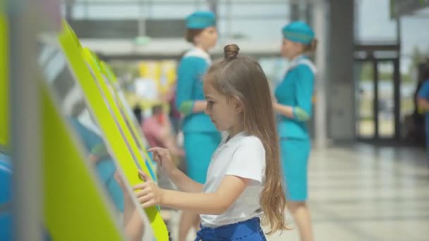 Vue latérale d'une petite fille concentrée utilisant un terminal de paiement à l'aéroport avec des hôtesses floues parlant en arrière-plan. mignon caucasien enfant en attente pour départ dans terminal. — Video