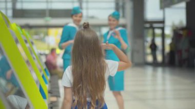 Havaalanındaki güzel hostesleri selamlayan küçük bir kızın arka görüntüsü. Kabin görevlisi üniformalı gülümseyen beyaz kadınlara odaklanın. Esmer çocuğa selam verin. Kalkışı bekliyor.