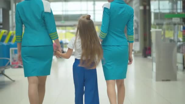 Visszapillantás a boldog kaukázusi lányra, aki profi légikísérők kezét fogja, és a reptéri kijárat felé sétál. Barna gyermek sétál a légiutas-kísérőkkel érkezés után. A felügyelet koncepciója. — Stock videók