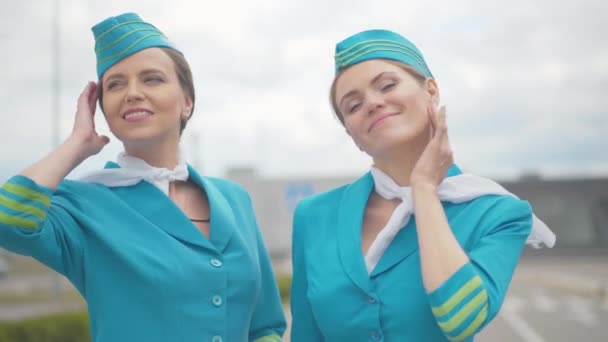 Middle shot of two confident women in flight attendant uniform standing outdoors on windy and and looking at camera. Portrait of beautiful Caucasian stewardesses posing on runway in airport. — Stock Video