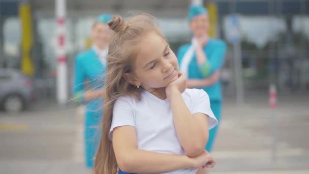 Retrato de una niña de ensueño de pie junto al aeropuerto con azafatas borrosas en el fondo y pensando. Encantadora morena Caucásico niño soñando con ser azafata. — Vídeos de Stock