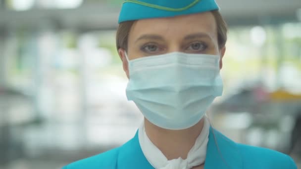 Close-up portret van serieuze blanke stewardess in gezichtsmasker kijkend naar camera. Professionele vrouwelijke stewardess poseren op de luchthaven tijdens coronavirus pandemie. Covid-19 internationaal toerisme. — Stockvideo