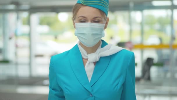 Fiatal stewardess egyenruhában és arcmaszkban a kamerába néz és mosolyog. Pozitív szakmai légiutas-kísérő portréja a Covid-19 világjárvány karantén repülőtéri terminálján. — Stock videók