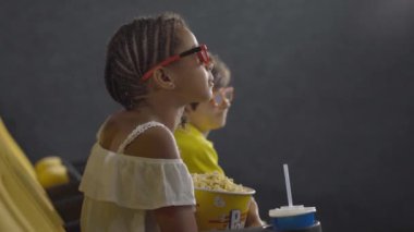 Arka planda bulanık çocuklar olan, sinemada 3D film izleyen küçük bir kızın yan görüntüsü. Üç boyutlu gözlüklü Afro-Amerikan çocuk, patlamış mısırla film izlerken şaşırmış yüz ifadesi yapıyor..