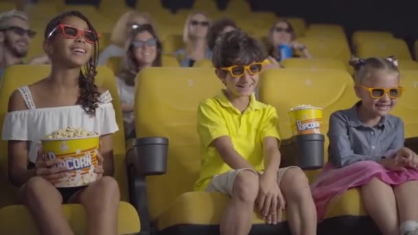 Gente multiétnica feliz en gafas 3D viendo comedia en el cine. Retrato de niños y adultos alegres disfrutando del cine en el cine. Niños positivos comiendo palomitas de maíz al frente y riendo. — Vídeo de stock