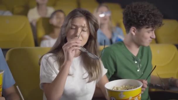 Glädjande ung kvinna sätta på 3D-glasögon och sitta ner på gul stol. Kameran rör sig mot självsäker vit man som tittar på film i biografen. Personer med popcorn njuter av 3D-film. — Stockvideo