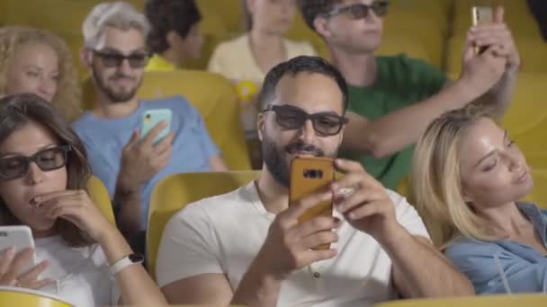 Visiteurs de cinéma joyeux utilisant des smartphones dans le cinéma. Portrait d'hommes et de femmes heureux et détendus de différentes ethnies en attente de film. Peuples caucasiens et du Moyen-Orient reposant à l'intérieur. — Video