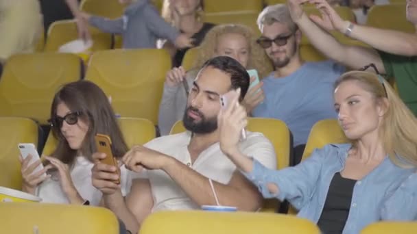 Homens e mulheres relaxados de diferentes etnias sentados com smartphones no cinema. Grupo de pessoas tirando selfie e navegando na Internet antes do cinema no cinema. — Vídeo de Stock