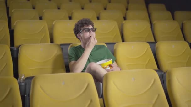 3d 안경을 쓴, 곱슬머리의 젊은 남자가 영화를 즐기는 넓은 샷. 영화 극장에서 혼자 앉아서 팝콘을 먹고 웃는 행복 한 남자. 무신경 한 영화 팬의 모습. — 비디오
