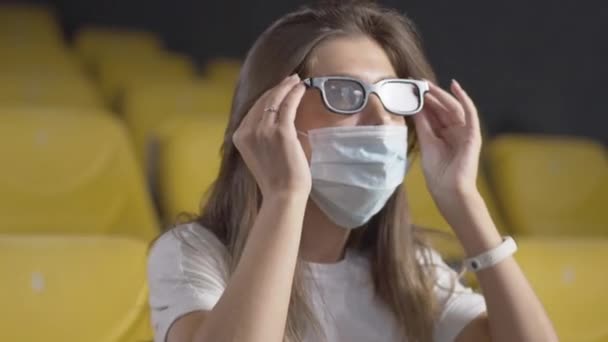 Primo piano della giovane bella donna bruna in maschera facciale che indossa occhiali 3d nel cinema. Ritratto di donna caucasica spensierata che guarda un film in sala cinematografica sulla pandemia di Covid-19 coronavirus. — Video Stock