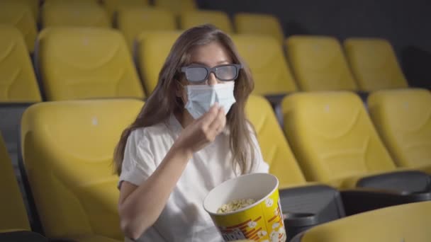 Retrato de una joven absorbida en mascarilla tratando de comer palomitas de maíz en el cine. Visitante caucásico absorto viendo películas en 3D en el cine durante el bloqueo pandémico del coronavirus. Estilo de vida Covid-19. — Vídeos de Stock