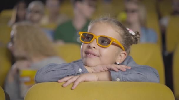 Charmante fille caucasienne absorbée regardant un film 3D au cinéma. Portrait d'un beau petit enfant dans des lunettes 3D assis sur une chaise jaune dans un cinéma et souriant. Visiteurs flous en arrière-plan. — Video