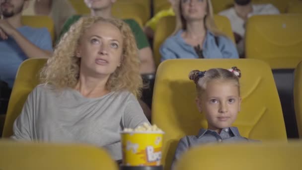 Utanmış Kafkasyalı anne sinemada kızlarının gözlerini kapatıyor. Sinema salonunda uygunsuz film izleyen rahat ebeveyn ve çocuk portresi. Boş zaman, ebeveynlik ve çocukluk. — Stok video