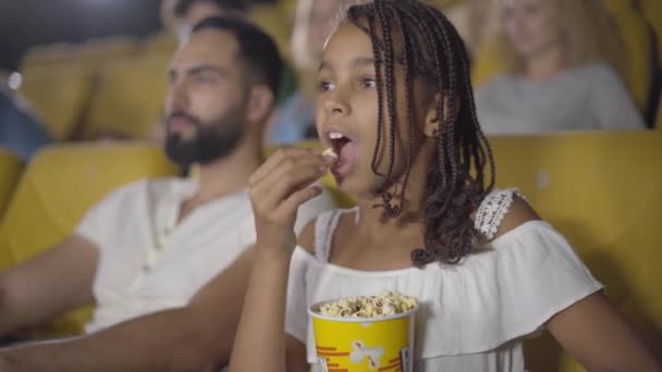Jolie fille afro-américaine mangeant du pop-corn au cinéma et montrant le pouce en l'air. Portrait d'enfant heureux avec des queues de cochon afro appréciant le cinéma. Enfant joyeux souriant à la caméra. — Video