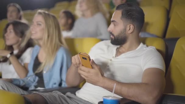 Handsome young Middle Eastern man using smartphone in cinema with annoying Caucasian women talking at the background. Portrait of irritated film-lover distracted from film in movie theatre. — Stock Video