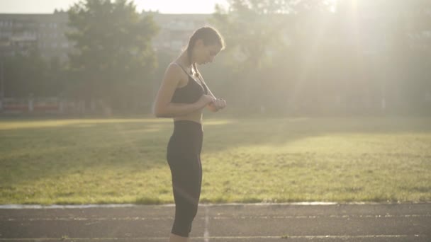 Slanke blanke vrouw lanceert app op slimme horloge en beginnen met rennen. Zijaanzicht portret van zelfverzekerde jonge sportvrouw training bij zonsopgang buiten. Mooie jogger controleert workout met toepassing. — Stockvideo