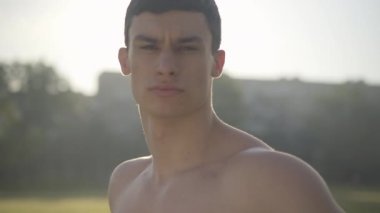 Güneş ışınlarındaki yakışıklı beyaz adamın yakın plan portresi. Yaz sabahında güneş ışığında poz veren ciddi sporcu. Kahverengi gözlü esmer adam kameraya bakıyor..