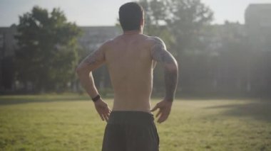 Dövmeli bir sporcunun güneş ışığında durup ellerini kalçalarına koyuşunun arkası. Genç, beyaz bir adam sabah antrenmanından sonra dışarıda dinleniyor. Kendine güvenen atletik adam antrenmandan zevk alıyor..
