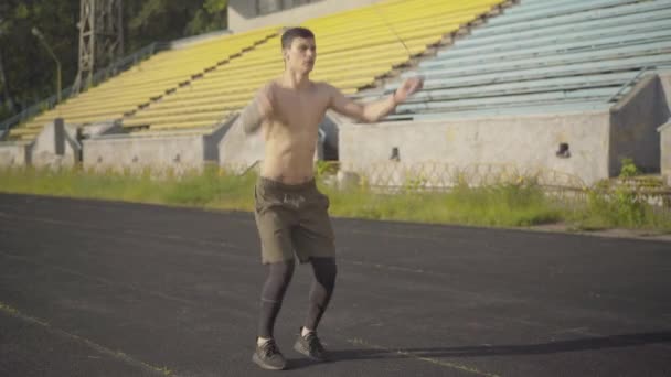 Concentrated sportsman jumping rope on stadium in summer morning. Portrait of Caucasian man exercising outdoors in sunlight. Wide shot of handsome sportive guy training at sunrise. — Stock Video