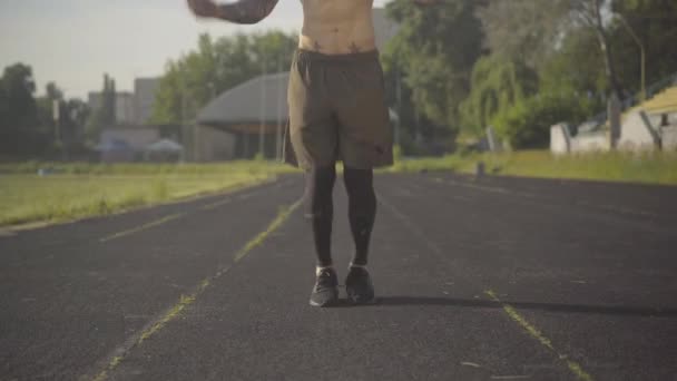 Vooraanzicht van de jonge Kaukasische sportman springtouw op sportveld. Portret van geconcentreerde mannentraining in de zomerochtend op het stadion. Zelfverzekerde man die traint in zonlicht. Camera beweegt omhoog. — Stockvideo