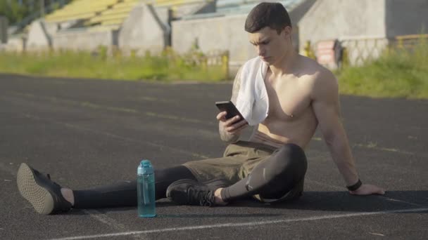 Сильный красивый спортсмен, сидящий на атлетическом поле и использующий смартфон. Портрет уверенного кавказца, занимающегося серфингом в Интернете после утренней тренировки на открытом воздухе. Парень отдыхает на стадионе на рассвете. — стоковое видео