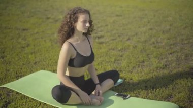 Gevşek kıvırcık saçlı kadın ellerini birleştiriyor ve nefes veriyor. Açık havada yaz sabahı egzersiz minderinde kendine güvenen beyaz yogi eğitiminin portresi. Meditasyon ve egzersiz kavramı.