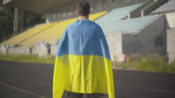 Rückenansicht eines selbstbewussten Sportlers, der die Hände mit ukrainischer Flagge auf dem leeren Stadion im Freien ausstreckt. Brünetter kaukasischer Mann steht im Sommermorgen mit Nationalsymbol auf dem Sportplatz. — Stockvideo