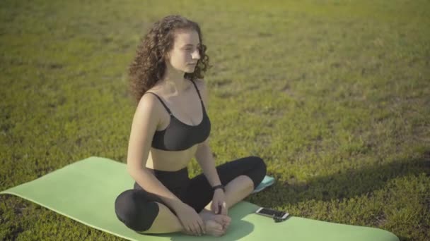 Donna dai capelli ricci rilassati che mette insieme le mani ed espira. Ritratto di allenamento caucasico sicuro di sé su un materassino da ginnastica in estate mattina all'aperto. Meditazione e concetto di allenamento. — Video Stock