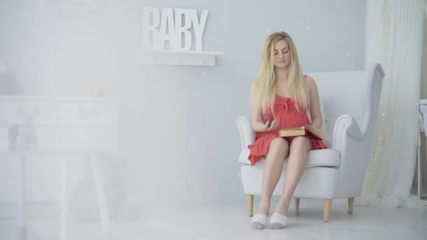 매력적 인 금발의 임산부가 책을 읽고 사과를 먹는 장면이요. 집안에서 임신을 즐기고 있는 아름다운 백인 여성의 행복 한 모습. 자신있는 여자가 아기 방에 앉아서 웃고 있는 모습. — 비디오