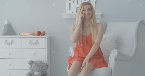 Cheerful pregnant woman sitting on armchair in baby room and talking on the phone. Portrait of beautiful young Caucasian expectant sharing news and smiling. Cinema 4k ProRes HQ. — Stock Video