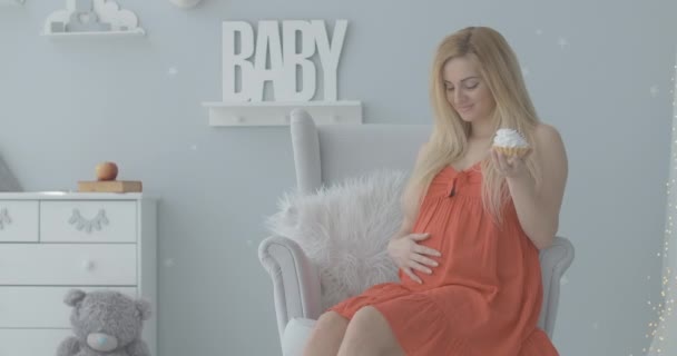 Happy young woman caressing pregnant belly and eating sweet creamy cake. Portrait of joyful Caucasian expectant enjoying tasty bun. Satisfied mother sitting in future baby room. Cinema 4k ProRes HQ. — Stock Video