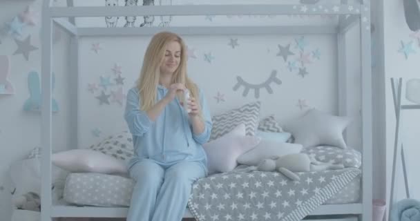 赤ん坊の部屋のベッドに坐り、健康なヨーグルトを飲む満足した妊婦。幸せなブロンド白人の期待の愛撫腹と笑顔の肖像。シネマ4k｜ProRes HQ. — ストック動画