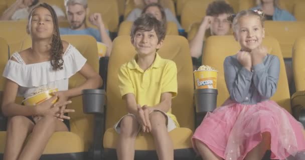 Drie opgewonden kinderen van verschillende etniciteiten die film kijken in de bioscoop met wazige mensen op de achtergrond. Portret van gelukkige kleine multi-etnische jongen en meisjes in de bioscoop. Cinema 4k ProRes Hoofdkwartier. — Stockvideo