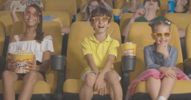 Sinemada 3D komedi izleyen mutlu çoklu etnik çocukların portresi. Arka planda bulanık yetişkinlerle gülen ve patlamış mısır yiyen 3 boyutlu gözlüklü tatlı çocuklar. Sinema 4k ProRes Merkezi.