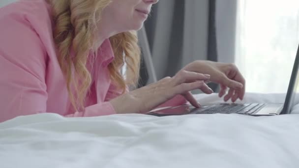 ベッドの上に横たわるノートパソコンのキーボードを入力する認識できない笑顔のシニア女性。自宅からオンラインで働く正の中高年白人のフリーランスの肖像画。フリーランスとリモートワークの概念. — ストック動画
