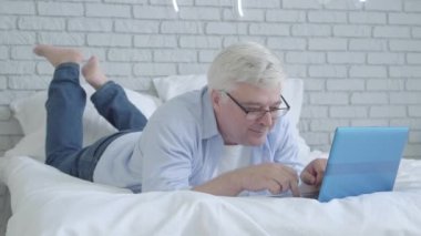 Yatak odasında dizüstü bilgisayarda gülümseyen son sınıf öğrencisi. Çevrimiçi, pozitif, orta yaşlı, Kafkasyalı emekli mesajlaşmasının portresi. İnternette sörf yapan mutlu aktif bir adam. Yatakta yatıyor..