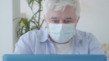 Covid-19 salgınında dizüstü bilgisayar kullanarak maskeli ciddi bir kıdemli adamın yakın çekimi. Coronavirus karantinası üzerine evden çalışan konsantre olmuş Kafkas orta yaşlı serbest çalışanın portresi.