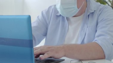 Covid-19 'daki yüz maskesi mesajında tanınmayan kıdemli serbest çalışan. Laptop klavyesinde yazı yazan orta yaşlı beyaz bir adam. Olgun iş adamı evde koronavirüs salgını üzerinde çalışıyor..